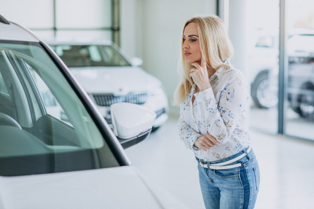 mulher loira olhando para um carro em uma loja de carros com expressão de duvida