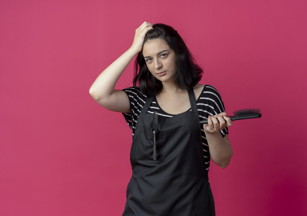 young-beautiful-female-hairdresser-apron-holding-hair-brush-confused-standing-pink-wall